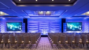 Un salón de conferencias con filas de sillas orientadas hacia una pantalla grande, un atril y un techo con iluminación azul.