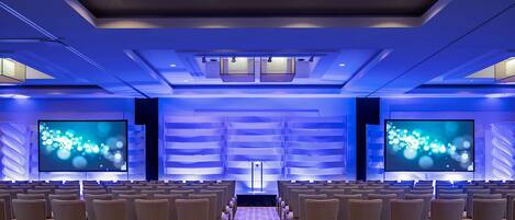 A conference room with rows of chairs facing a large screen, a podium, and a blue-lit ceiling.