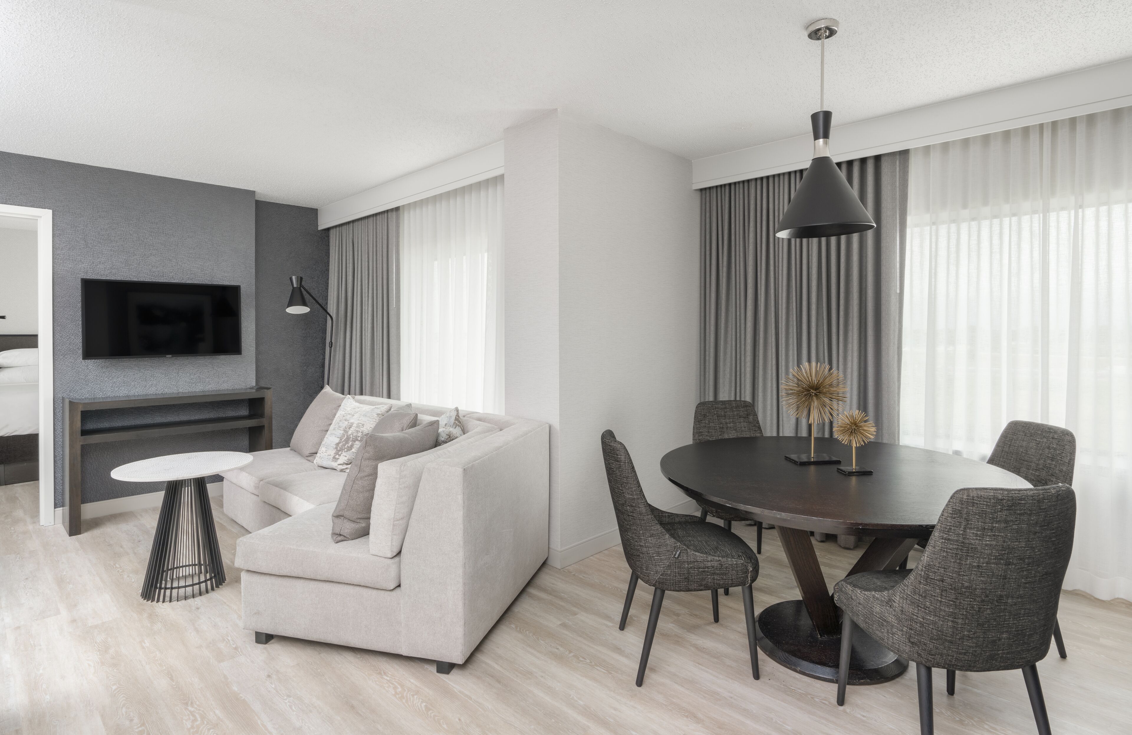 A modern living room with a sofa, a round dining table, and chairs. There is a TV mounted on the wall and a lamp on a side table.