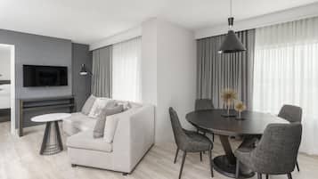 Una sala de estar moderna con un sofá, una mesa de comedor redonda y sillas. Hay un televisor montado en la pared y una lámpara en una mesita auxiliar.