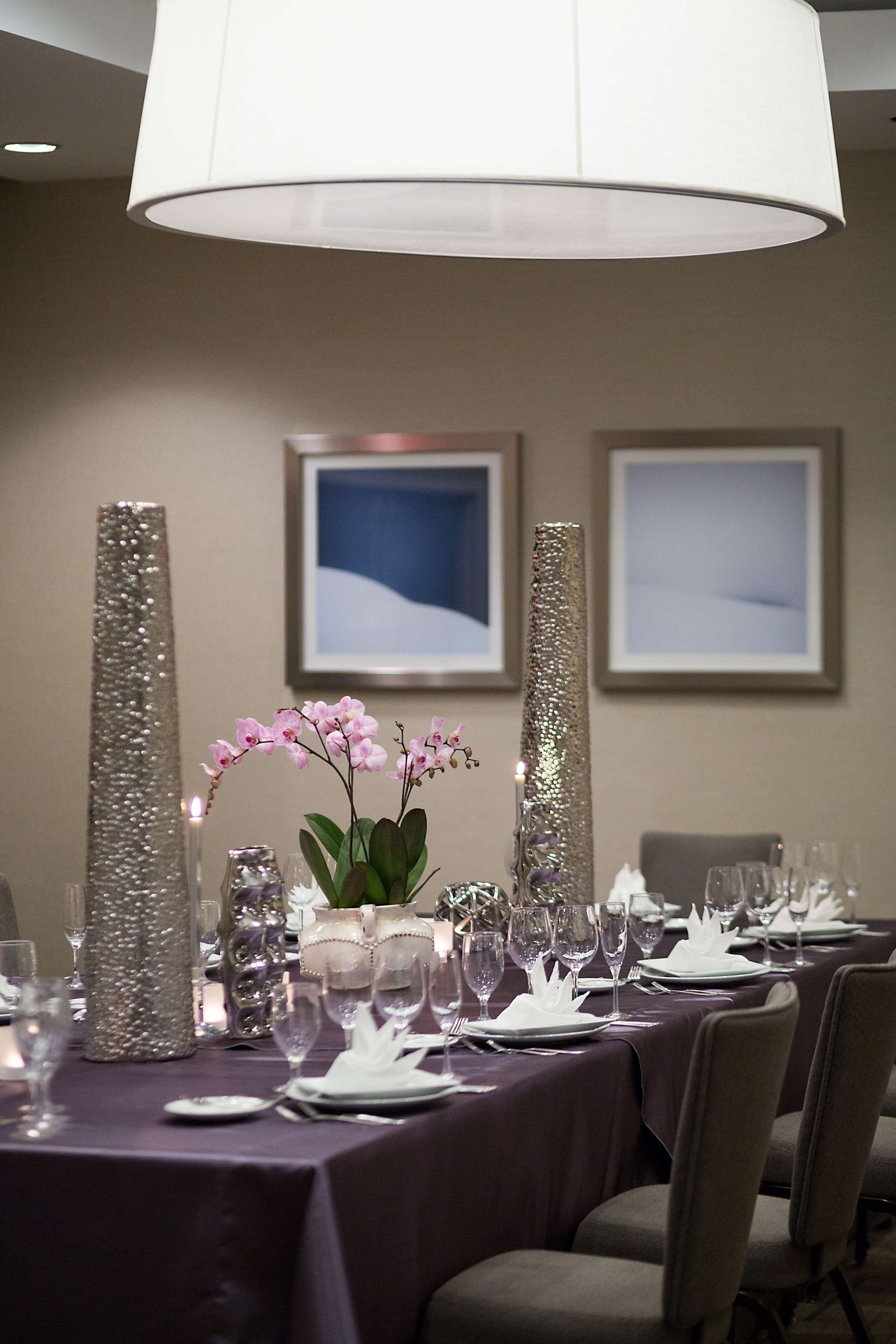 A dining table set with white plates, silverware, and glasses, flanked by tall, glittery vases and a vase with pink orchids.
