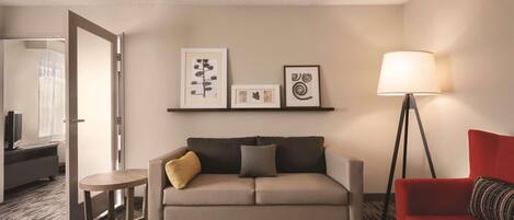 A hotel room with a sofa, a red armchair, a small table, and a floor lamp. There are framed pictures on the wall.