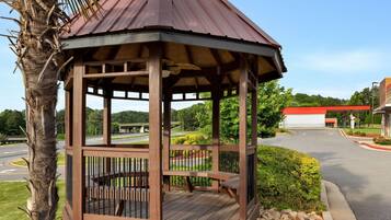 A wooden gazebo with a metal roof, surrounded by a landscaped garden and a road.