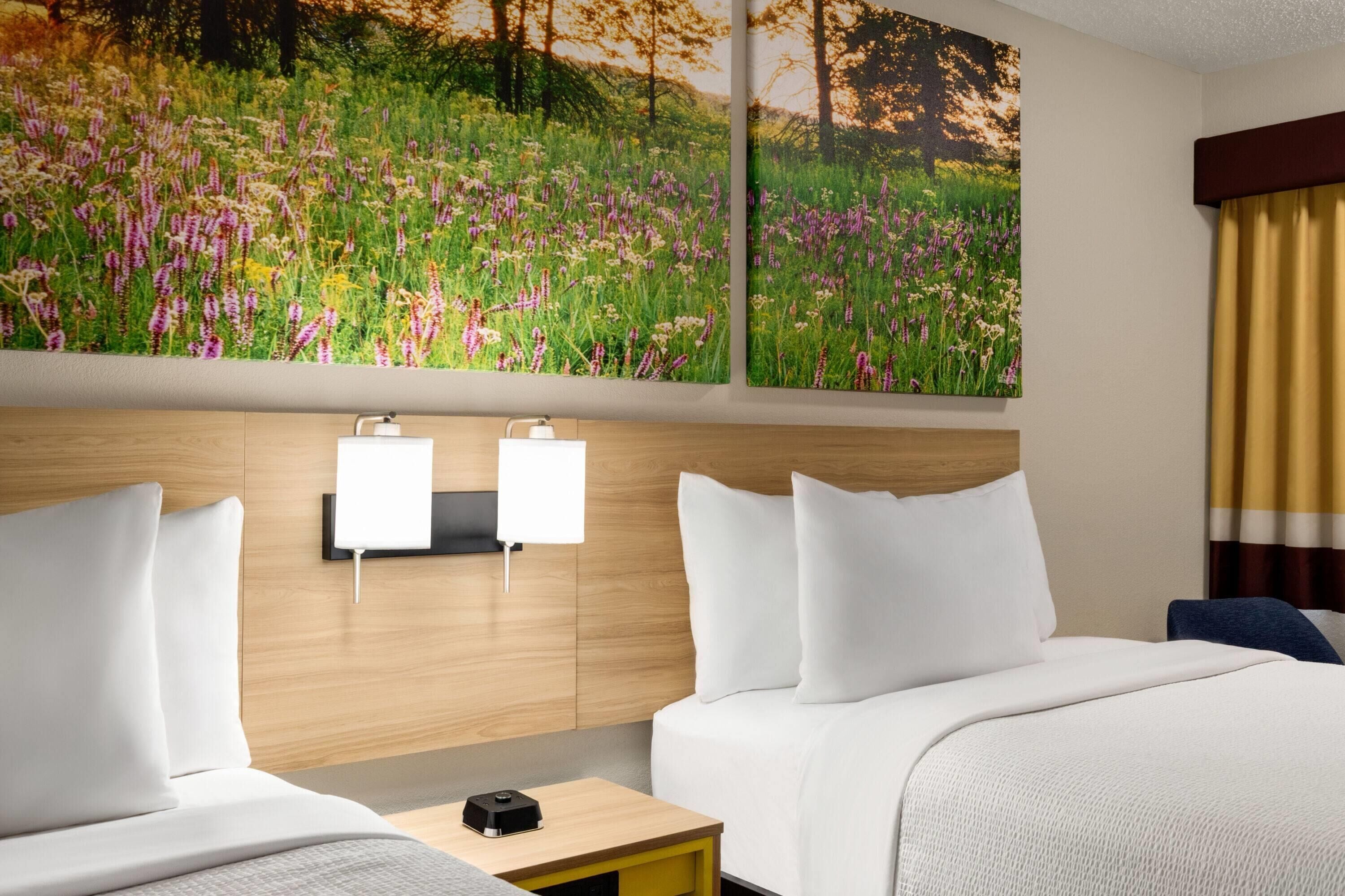 A hotel room with two beds, a nightstand, a wall-mounted lamp, and a large floral painting above the beds.