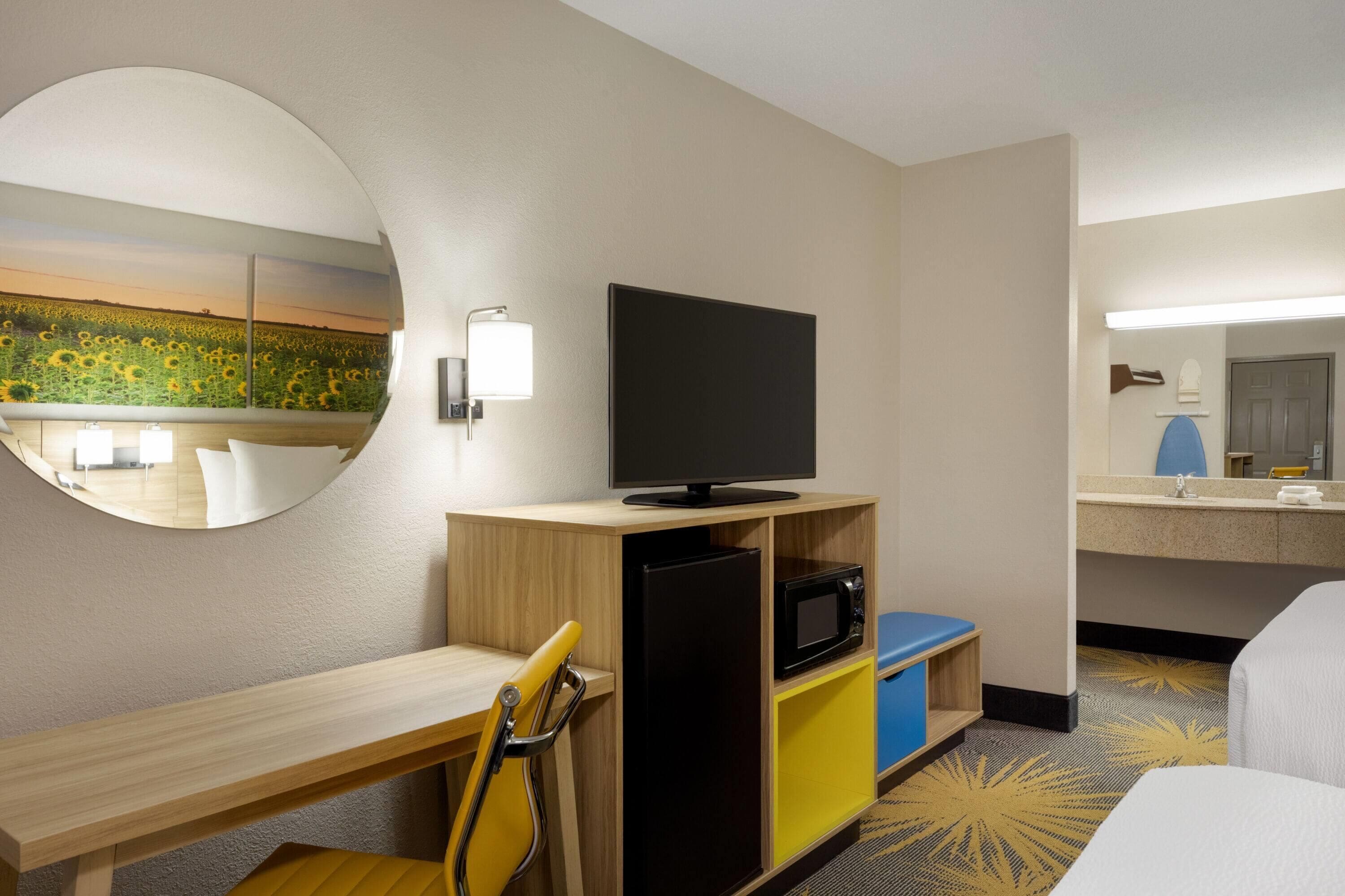 Une chambre d’hôtel équipée d’un bureau, d’une télévision, d’un four à micro-ondes, d’un petit réfrigérateur, d’une chaise, d’un miroir et d’une salle de bain avec un lavabo et un miroir.