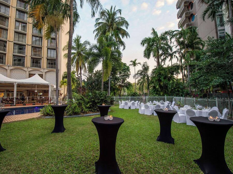 An outdoor event space with tables and chairs, surrounded by palm trees and a building in the background.