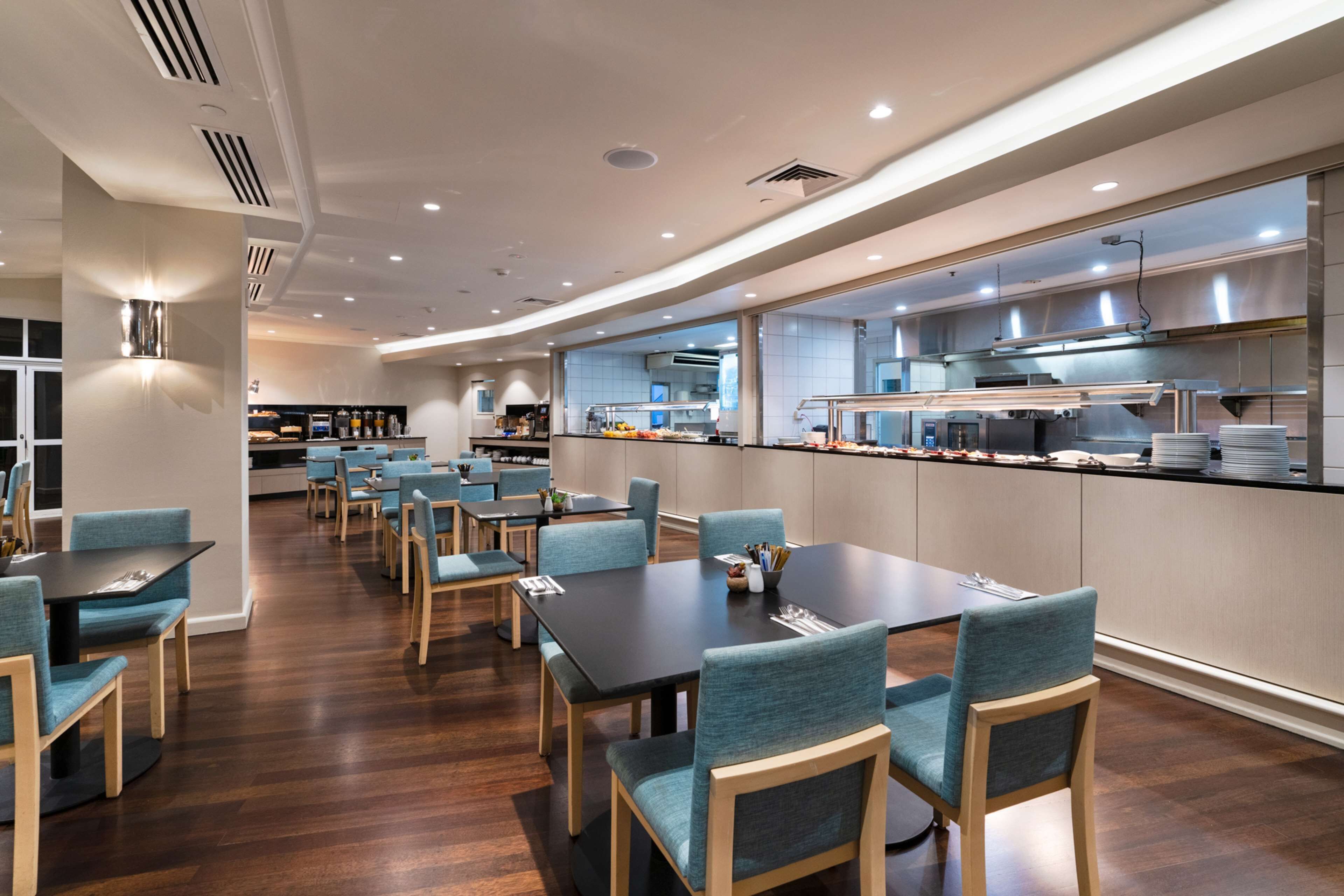 A modern dining area with multiple tables and chairs, a kitchen in the background, and a clean, well-lit environment.