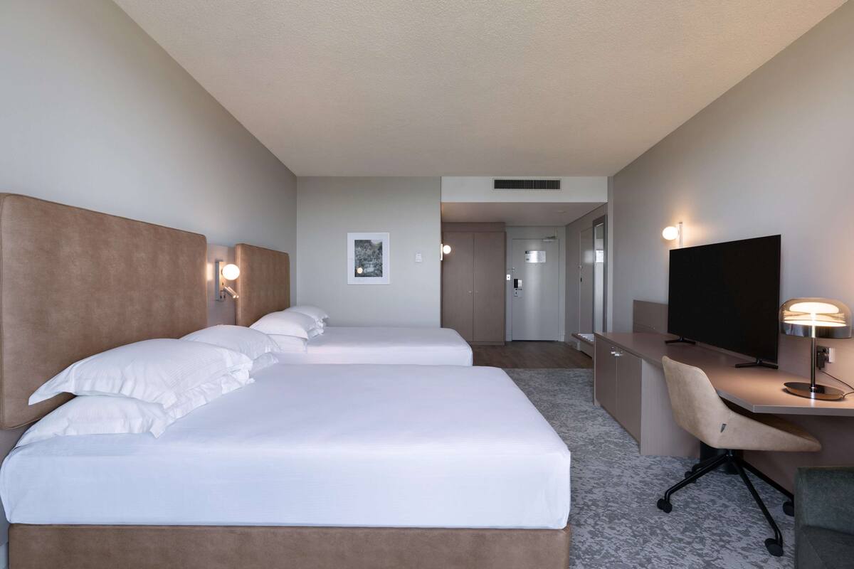 Habitación de hotel con dos camas, un escritorio, una silla, un televisor y una lámpara en la pared.