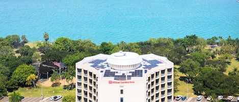 A Hilton Garden Inn hotel with a central courtyard, surrounded by parking and greenery, overlooking a body of water.
