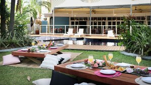 An outdoor dining area with tables set for a meal, a pool in the background, and a building with a balcony.