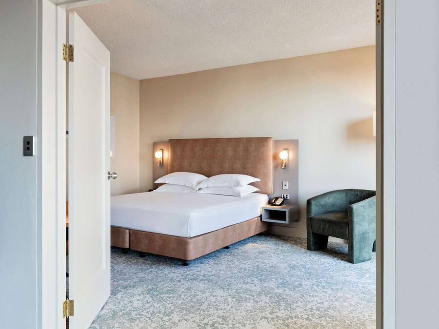 Una habitación de hotel con cama, mesita de noche, silla y una lámpara de pared.