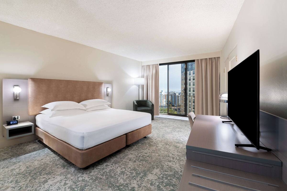Una habitación de hotel moderna con una cama grande, un escritorio, un televisor de pantalla plana y vistas a la ciudad a través de amplios ventanales.