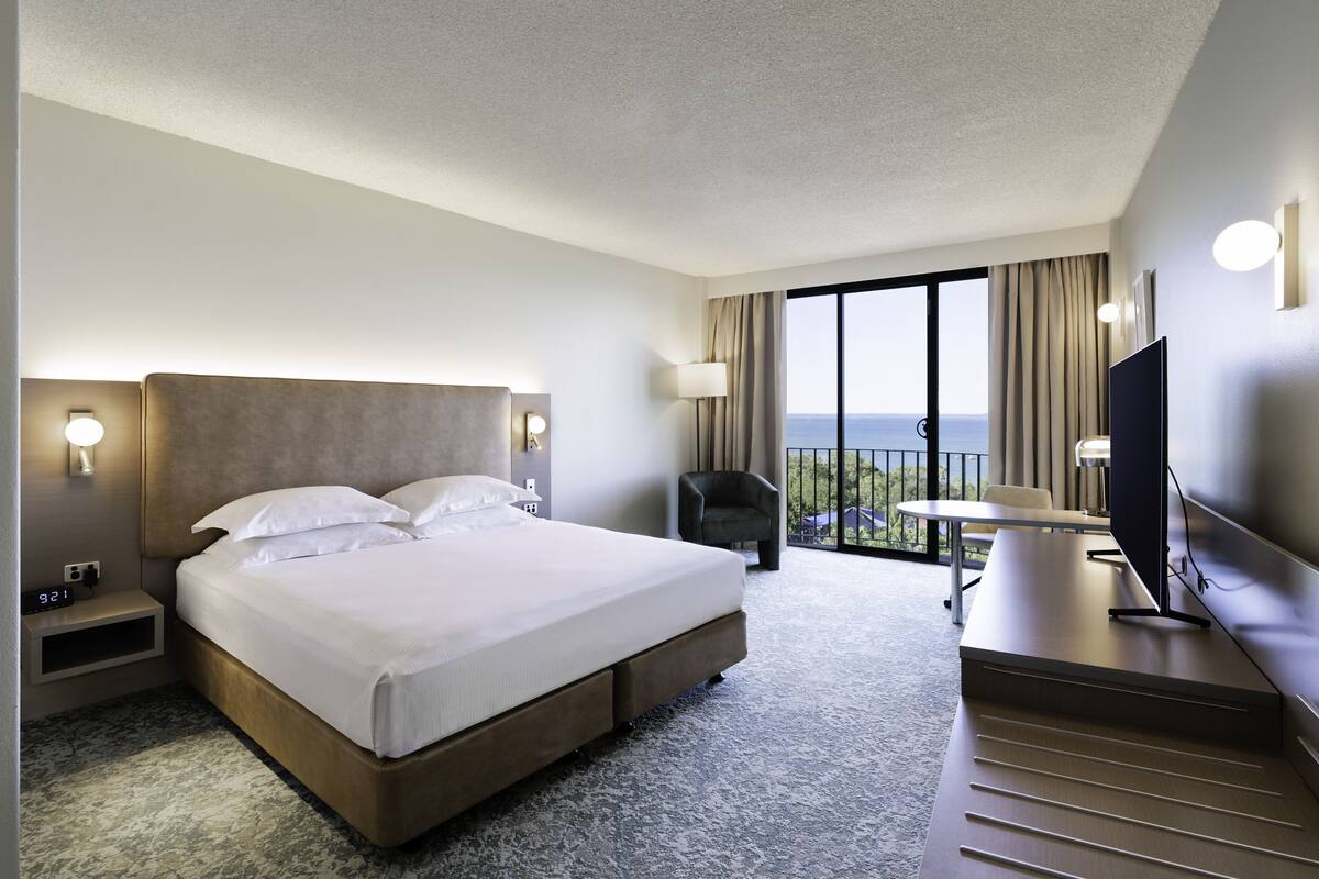 Habitación de hotel con una cama grande, un escritorio, una silla, un televisor y un balcón con vistas.