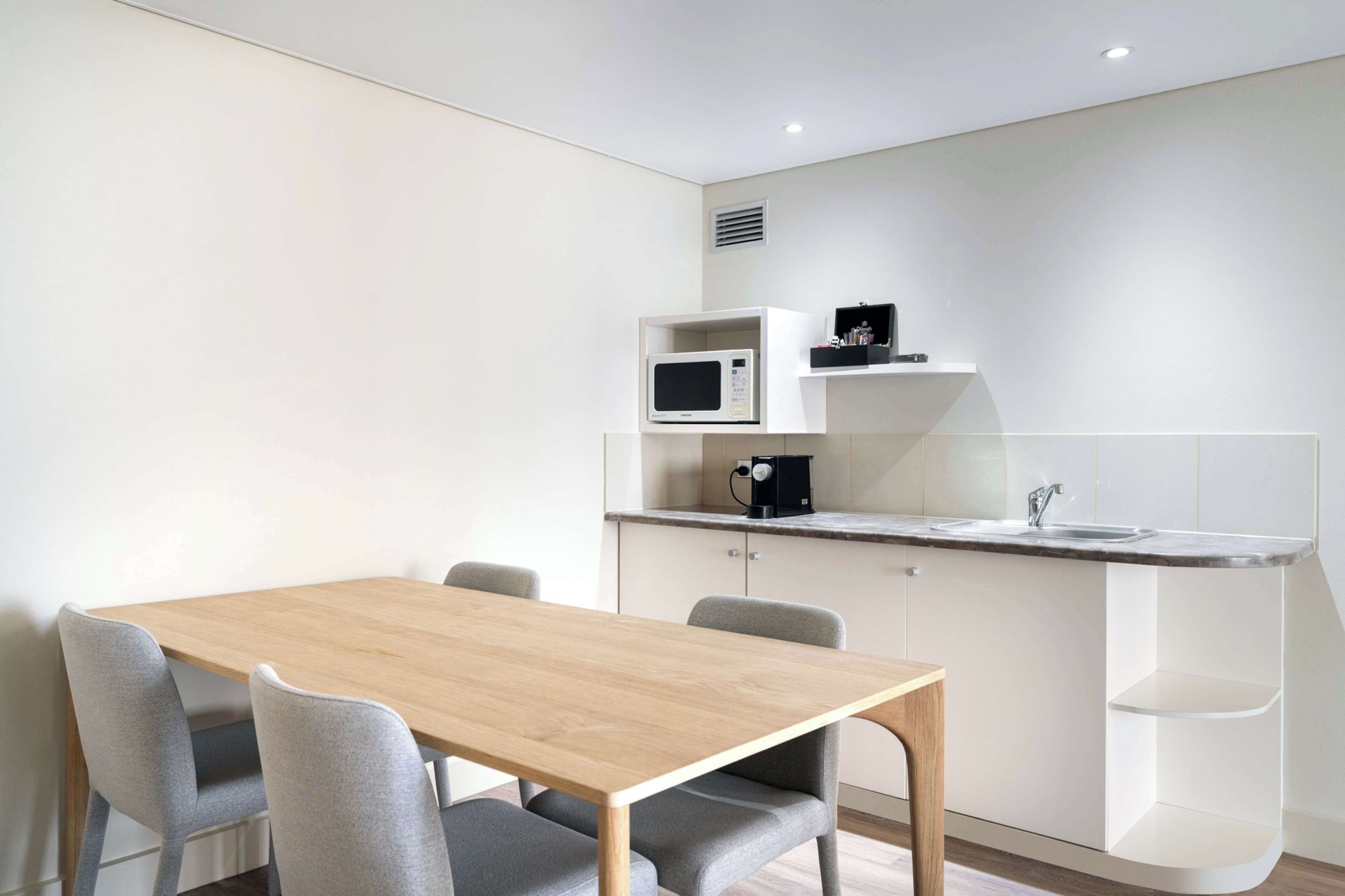 A modern kitchen with a wooden table and grey chairs, a microwave, a coffee machine, and a sink.
