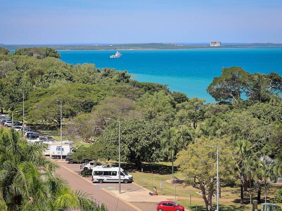 Vista costera con puerto deportivo, vehículos estacionados y abundante vegetación.