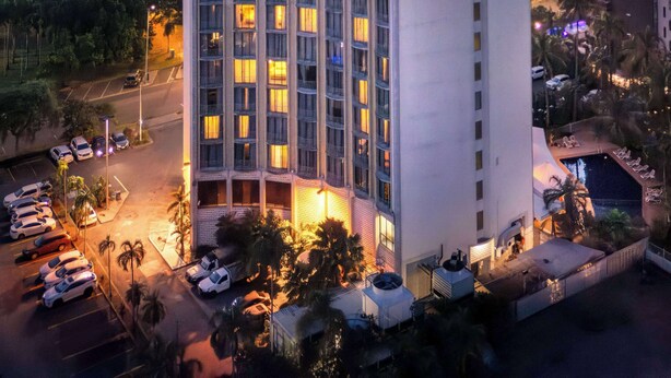 A high-rise hotel with multiple floors, a parking area with several cars, and a view of the sea at sunset.