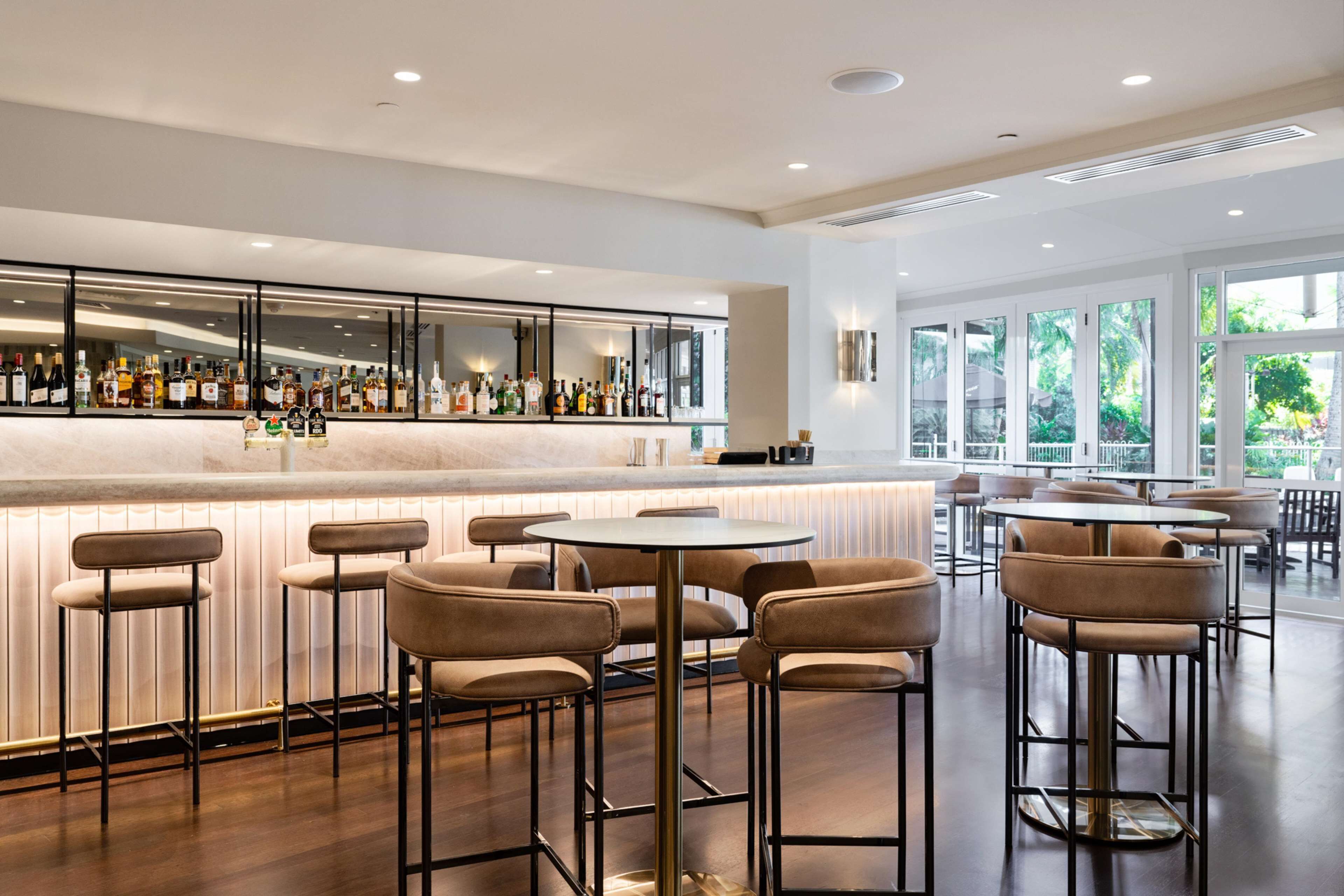 A modern bar area with a long counter displaying various bottles, surrounded by high stools and round tables.