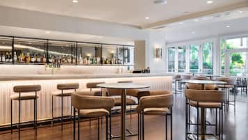 A modern bar area with a long counter displaying various bottles, surrounded by high stools and round tables.