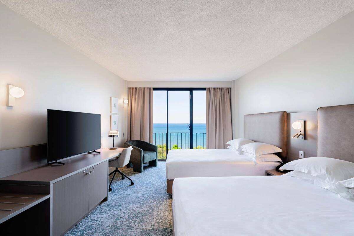 Habitación de hotel con dos camas, un escritorio con televisor, una silla y vista al océano.