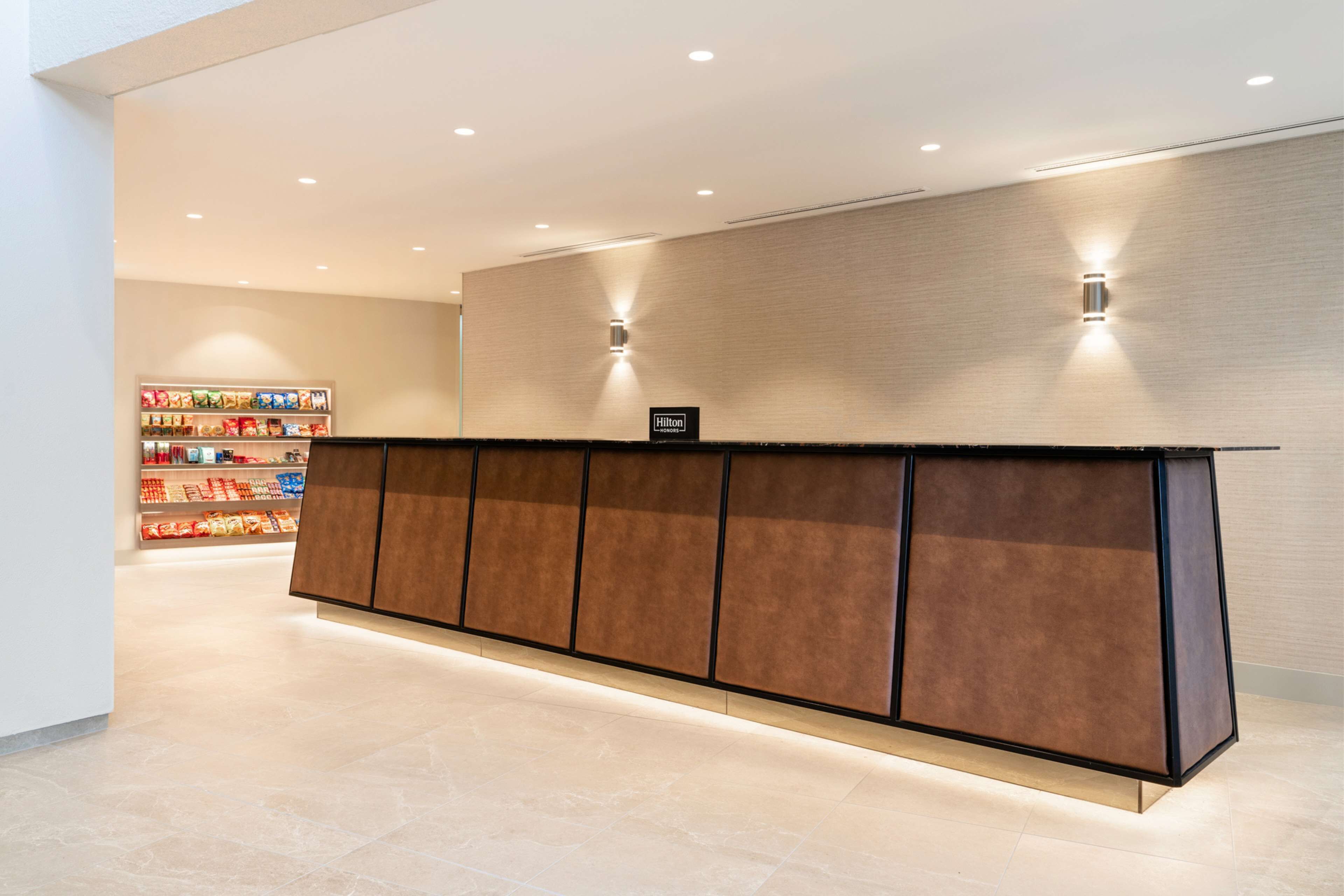 Ein Hotel-Rezeptionsbereich mit einer modernen Theke, einem Schild mit dem Markennamen und einer Snackauslage im Hintergrund.