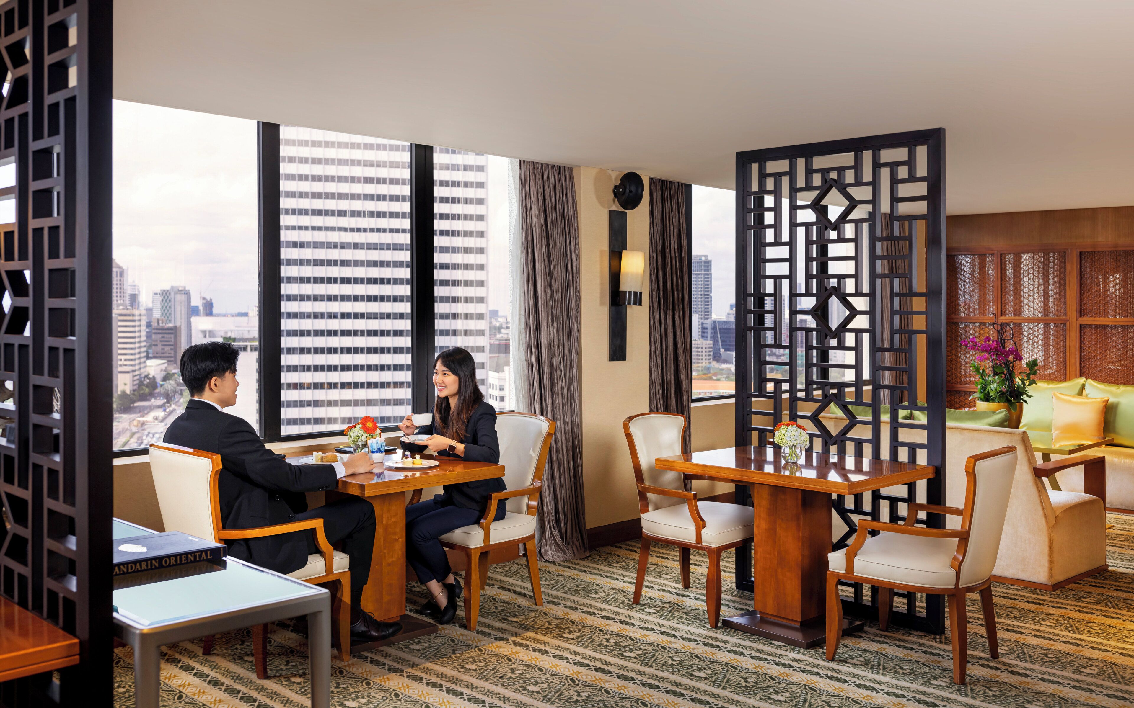 A hotel room with a large window offering a city view, a wooden dining table with chairs, and a decorative partition with a geometric pattern.