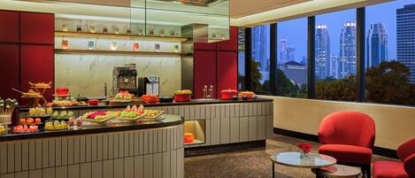 A modern hotel lobby with a kitchen area, a seating area with red chairs, and a view of the cityscape.