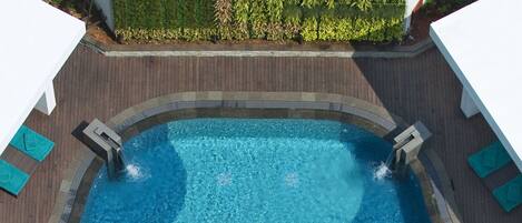 An aerial view of a swimming pool with a wooden deck, surrounded by white umbrellas and a green hedge.