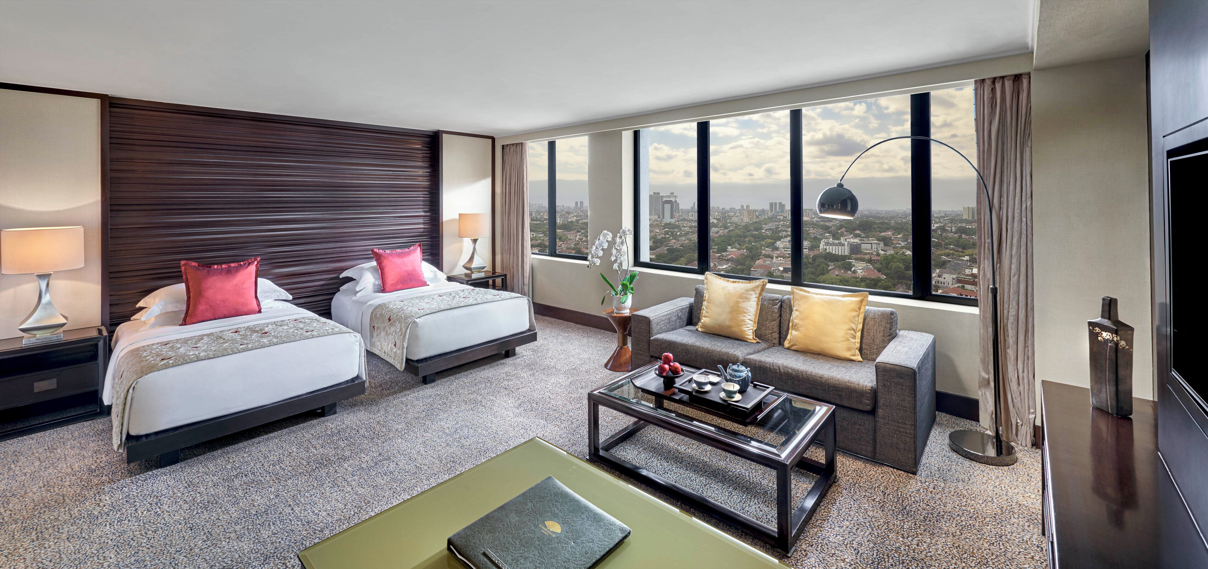 A hotel room with two beds, a sofa, a coffee table, and a TV. The room has a large window with a city view.