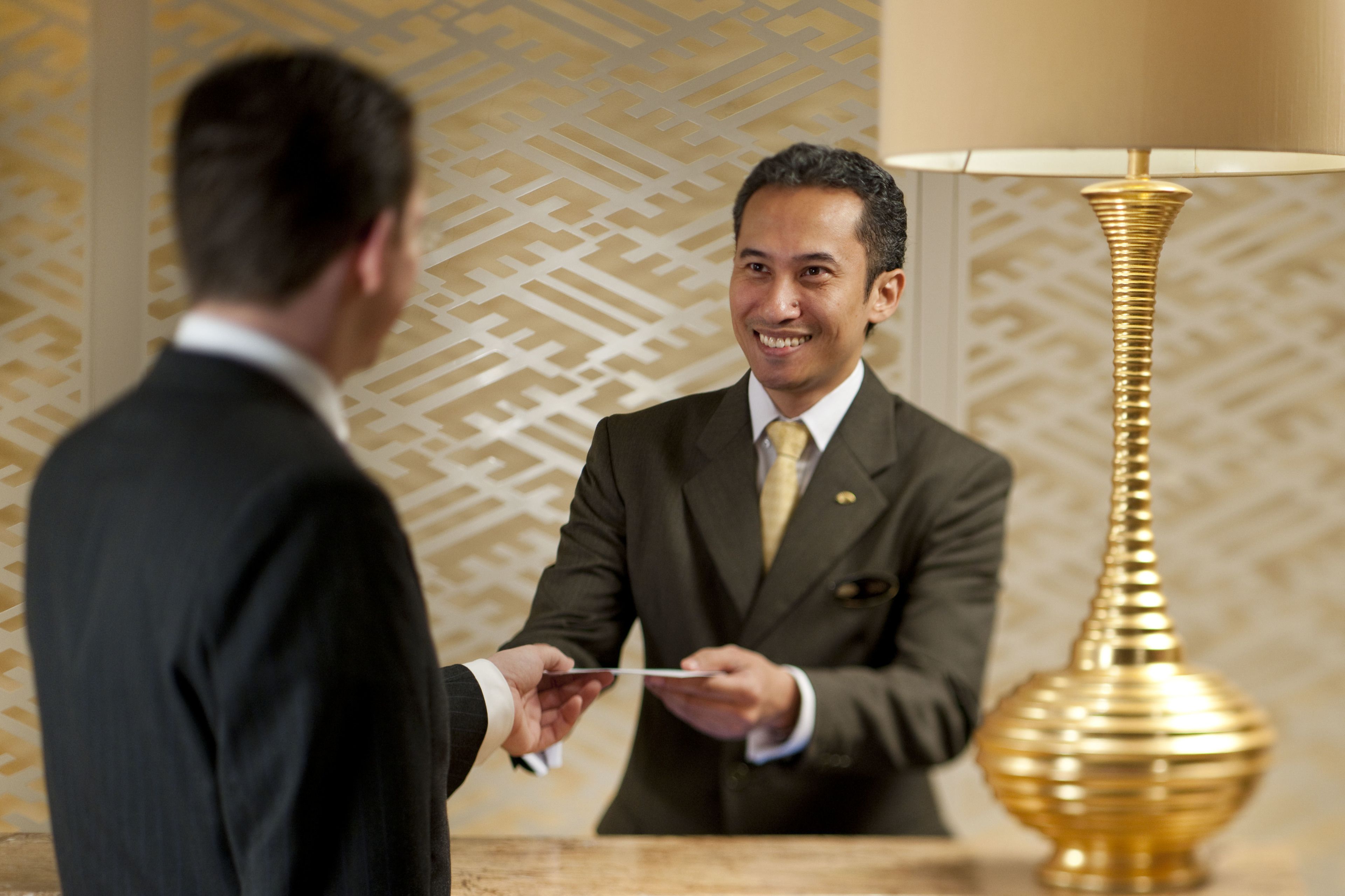 A man in a suit handing over a card to another man in a suit in a room with patterned wallpaper and a gold lamp.