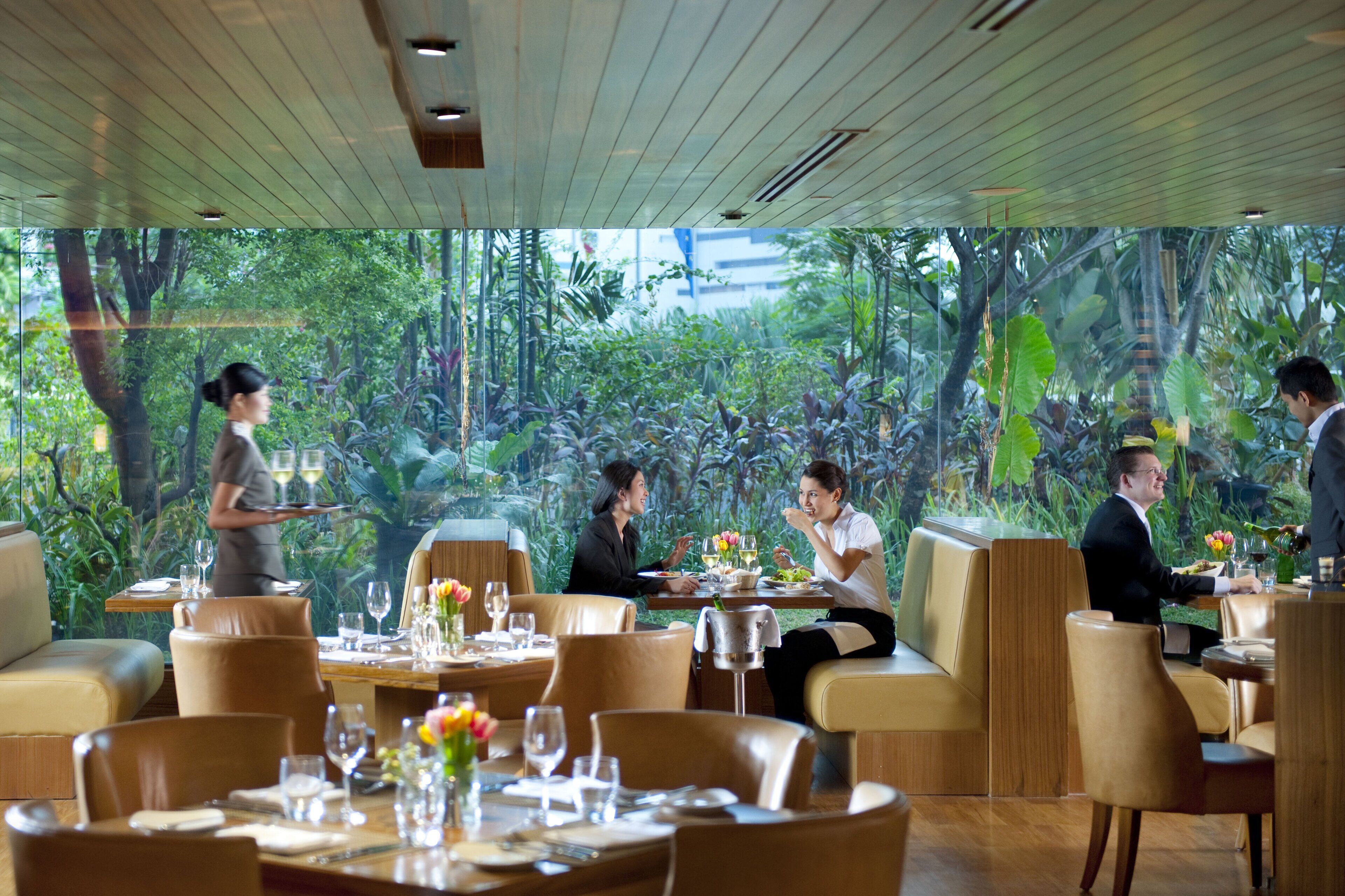 A restaurant with a view of lush greenery through large windows, leather seating, and a waiter attending to guests.