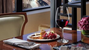 Una mesa de comedor con un plato de comida, una copa de vino tinto y un periódico. Una ventana muestra un paisaje urbano con edificios y una autopista al atardecer.