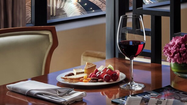Una mesa de comedor con un plato de comida, una copa de vino tinto y un periódico. Una ventana muestra un paisaje urbano con edificios y una autopista al atardecer.