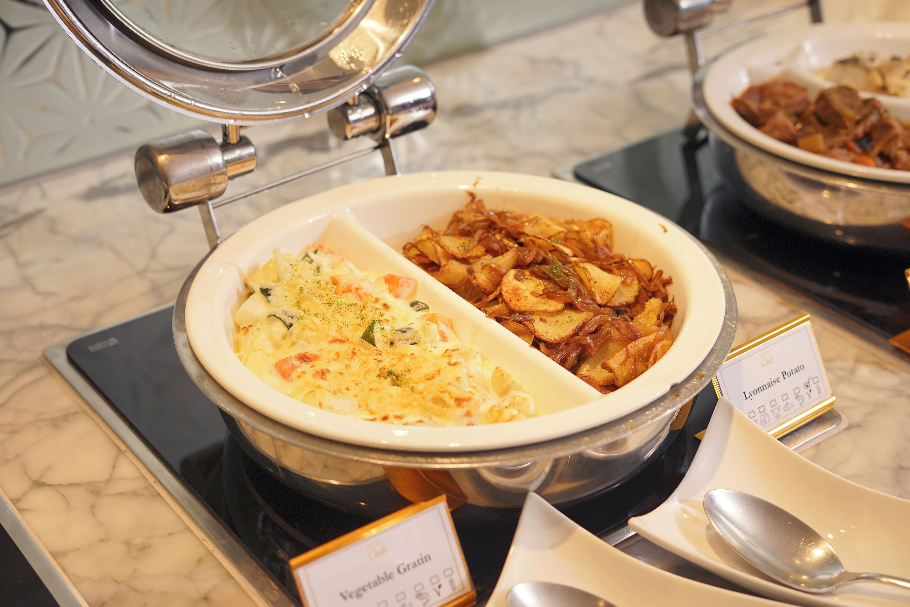A buffet with a variety of dishes, including a section labeled "Vegetable Gratin" and another labeled "Lyonnaise Potato."