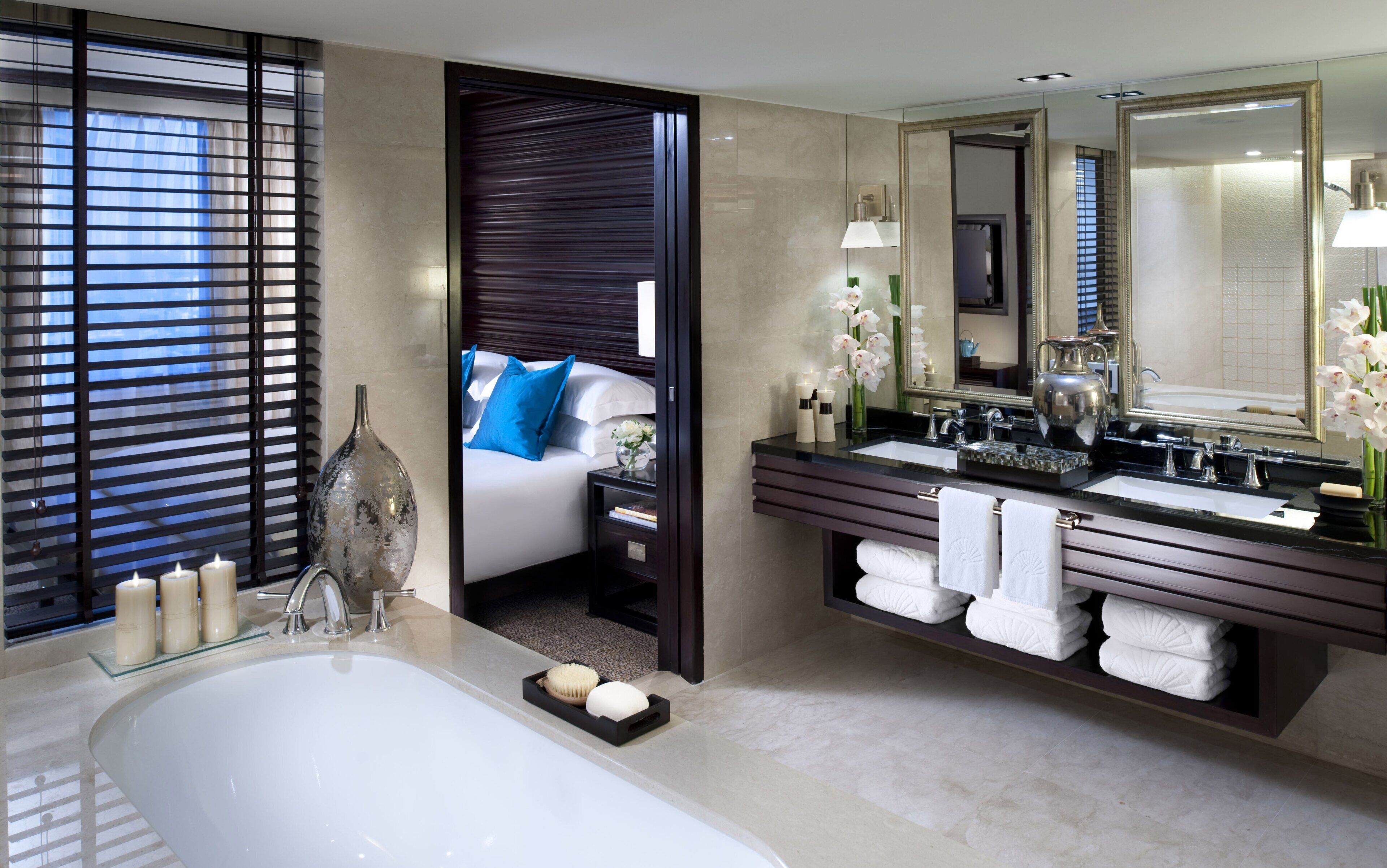 Un baño moderno con bañera, tocador doble con espejos, y una zona de dormitorio separada con cama y persianas.