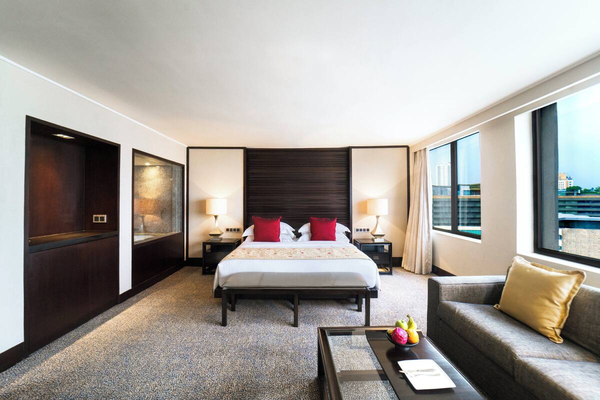 Habitación de hotel con una cama grande, dos mesitas de noche con lámparas, un sofá, una mesa de centro y amplias ventanas con vistas a la ciudad.