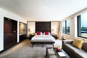 Habitación de hotel con una cama grande, dos mesitas de noche con lámparas, un sofá, una mesa de centro y amplias ventanas con vistas a la ciudad.