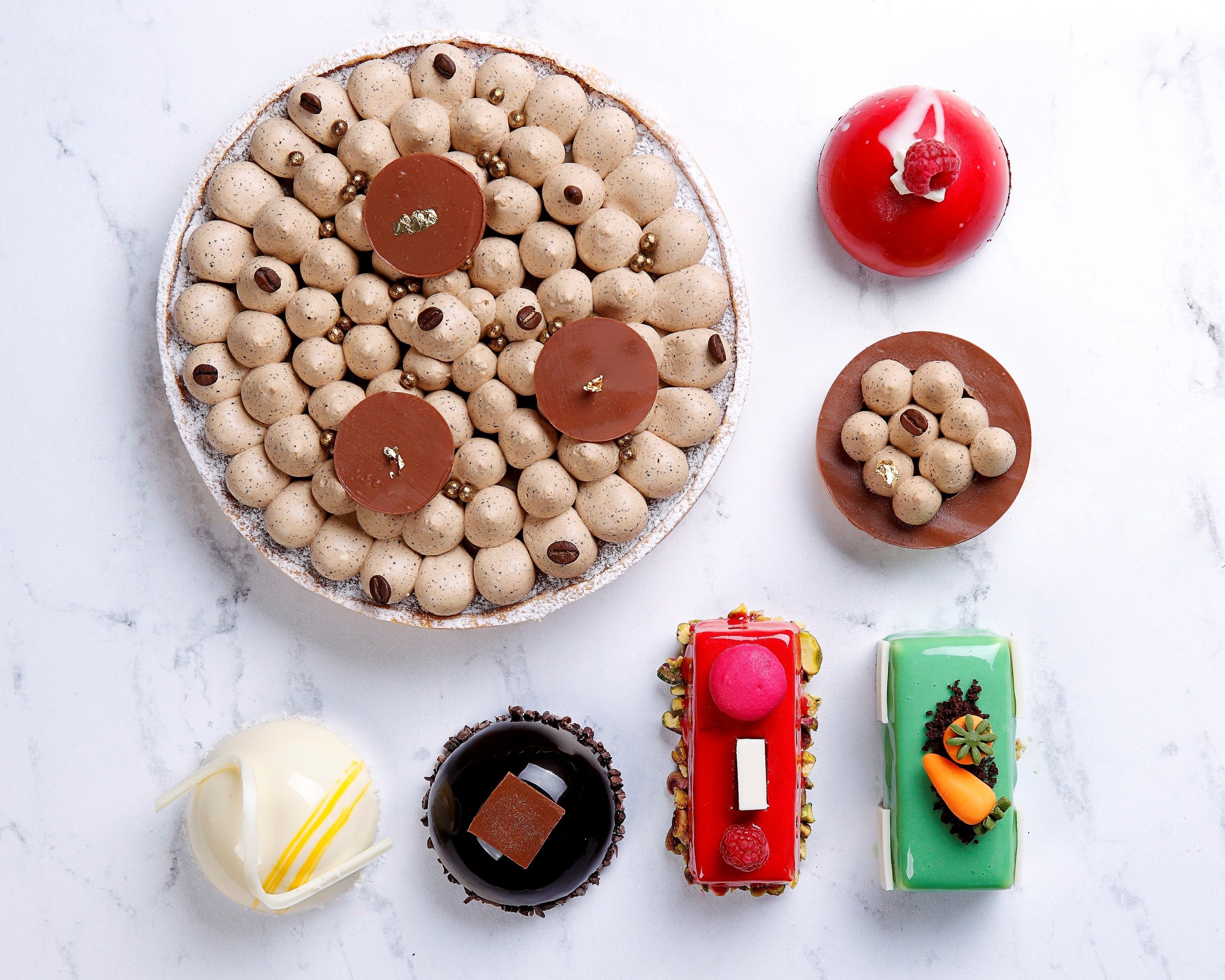 A variety of desserts including a large cake with chocolate and coffee beans, a red dessert with a raspberry, and smaller cakes with different toppings.