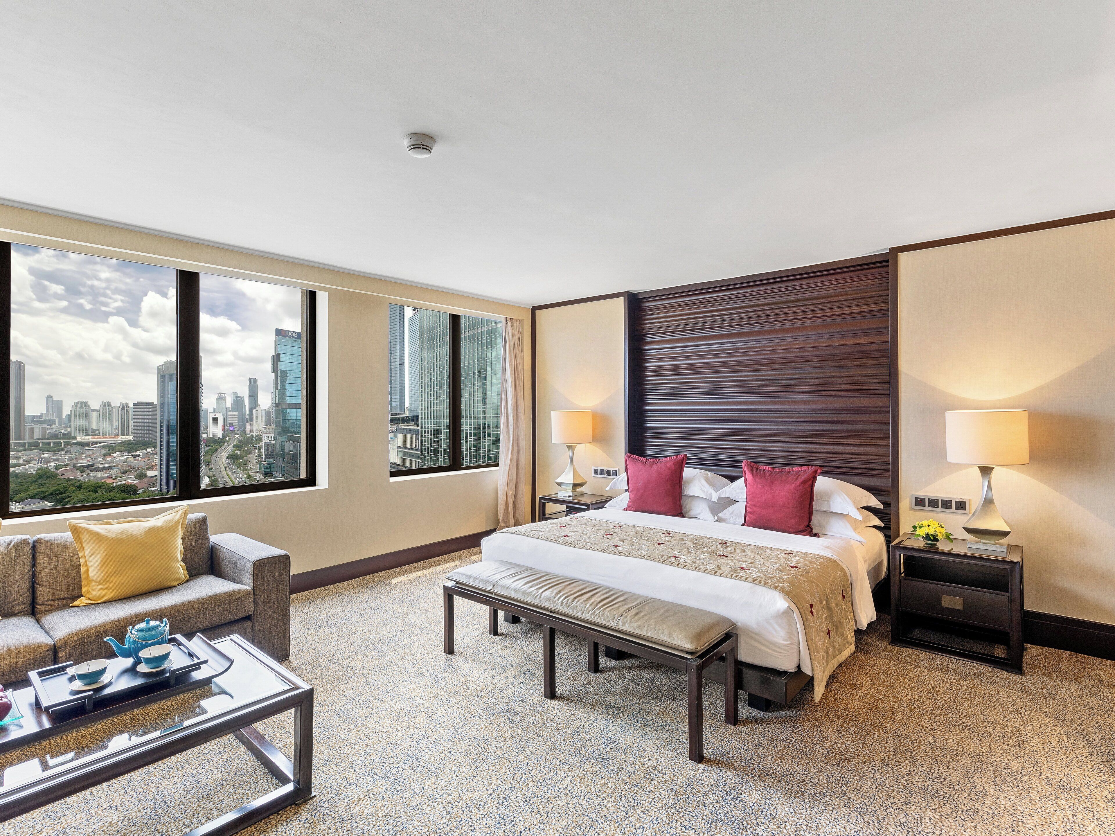 A hotel room with a large bed, two bedside tables with lamps, a sofa, a coffee table, and a city view through large windows.