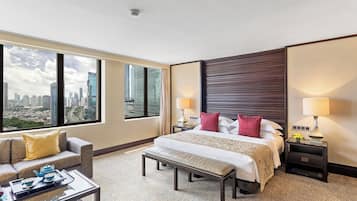 Habitación de hotel con una cama grande, dos mesitas de noche con lámparas, un sofá, una mesa de centro y vistas a la ciudad a través de amplios ventanales.