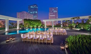 Un área de comedor al aire libre con piscina, rodeada de follaje y vistas a un paisaje urbano al atardecer.