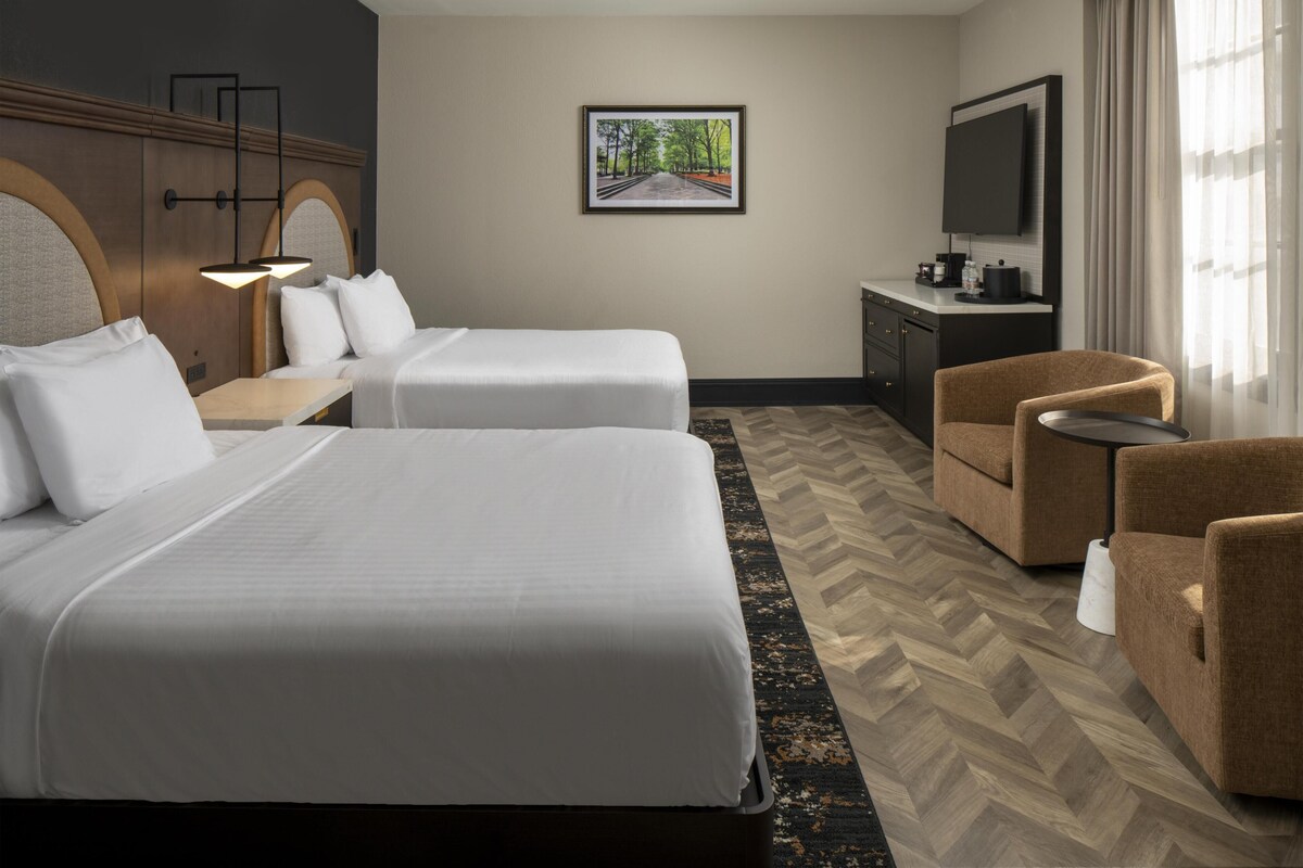 A hotel room with two beds, a desk, a chair, a TV, and a herringbone-patterned floor.