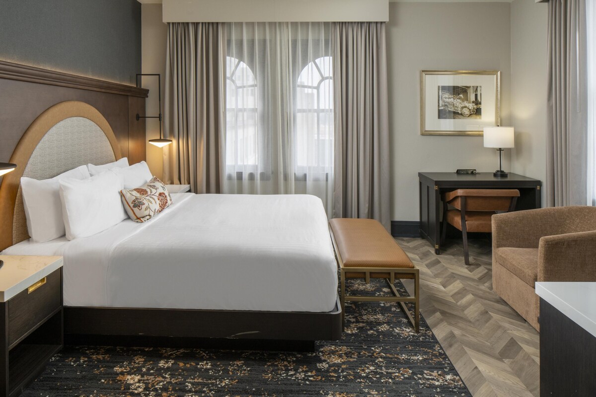 A hotel room with a large bed, a desk, a chair, and a bench.
