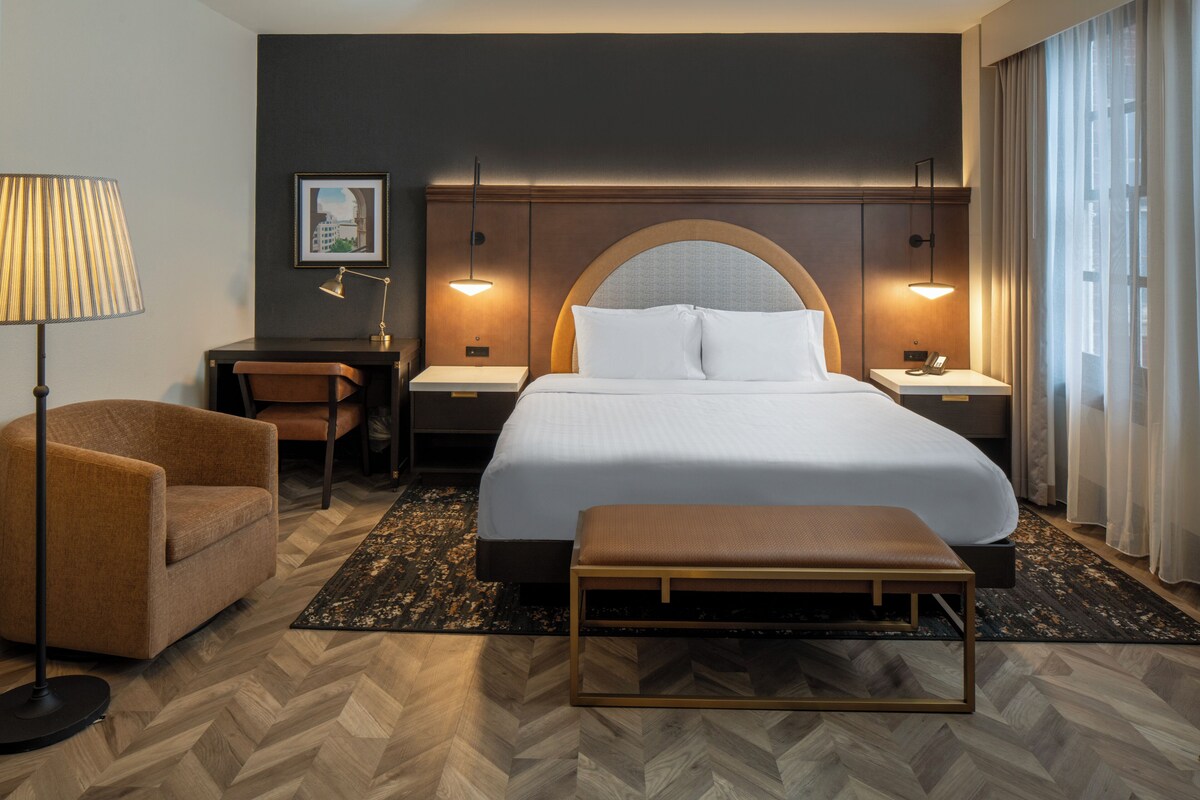 A hotel room with a bed, bedside tables, a lamp, a chair, and a bench.