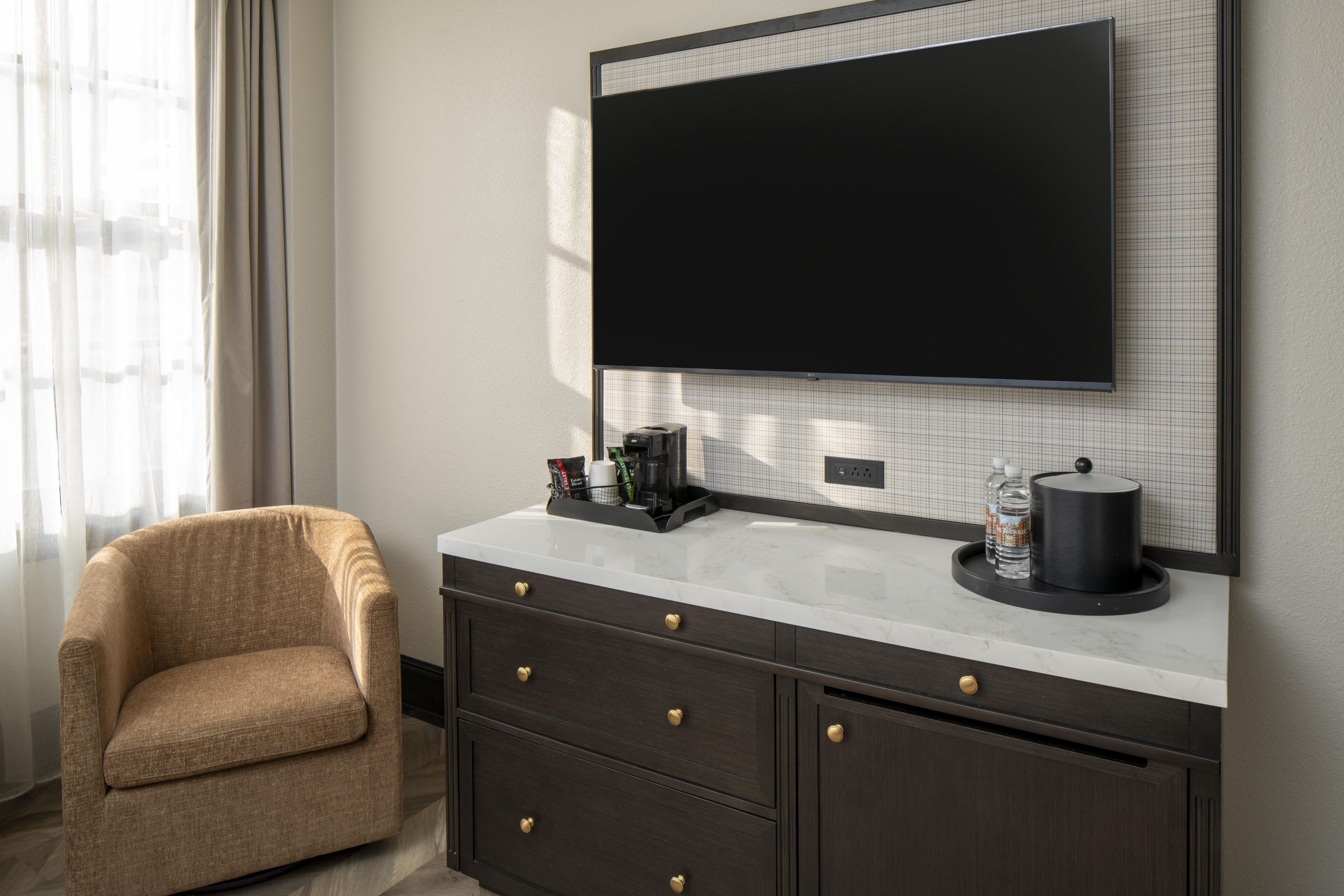 Una habitación de hotel moderna con una televisión de pantalla plana montada en la pared, un aparador de madera oscura con cajones, un sillón color beige y una ventana con cortinas translúcidas.