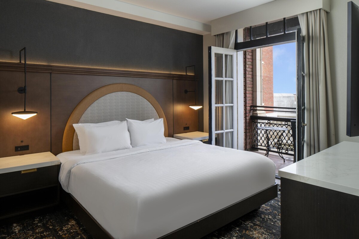 A hotel room with a large bed, two bedside tables, a headboard with a circular design, a small table, and a view of the outside.