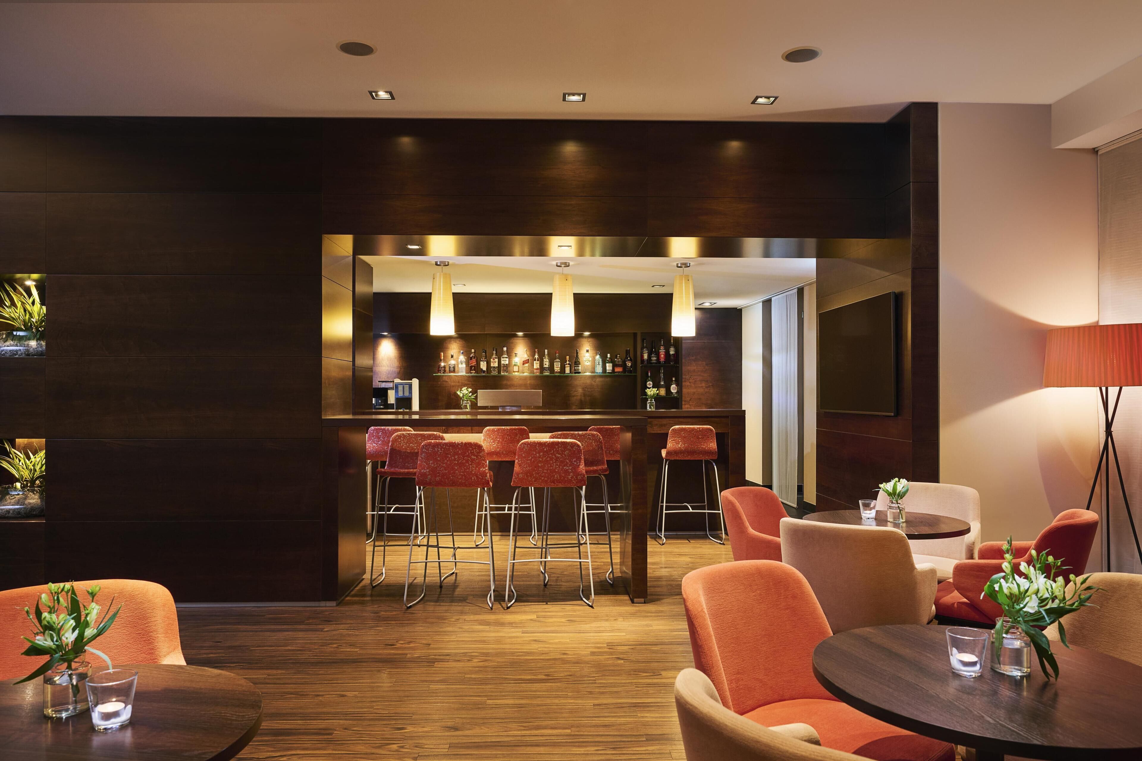 Zona bar moderna con pavimento in legno, sgabelli rossi e lampade a sospensione.