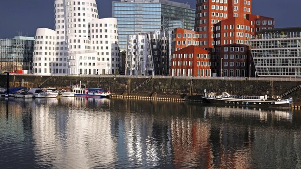 Paesaggio urbano con edifici moderni, un fiume tranquillo e barche ormeggiate sulla riva.