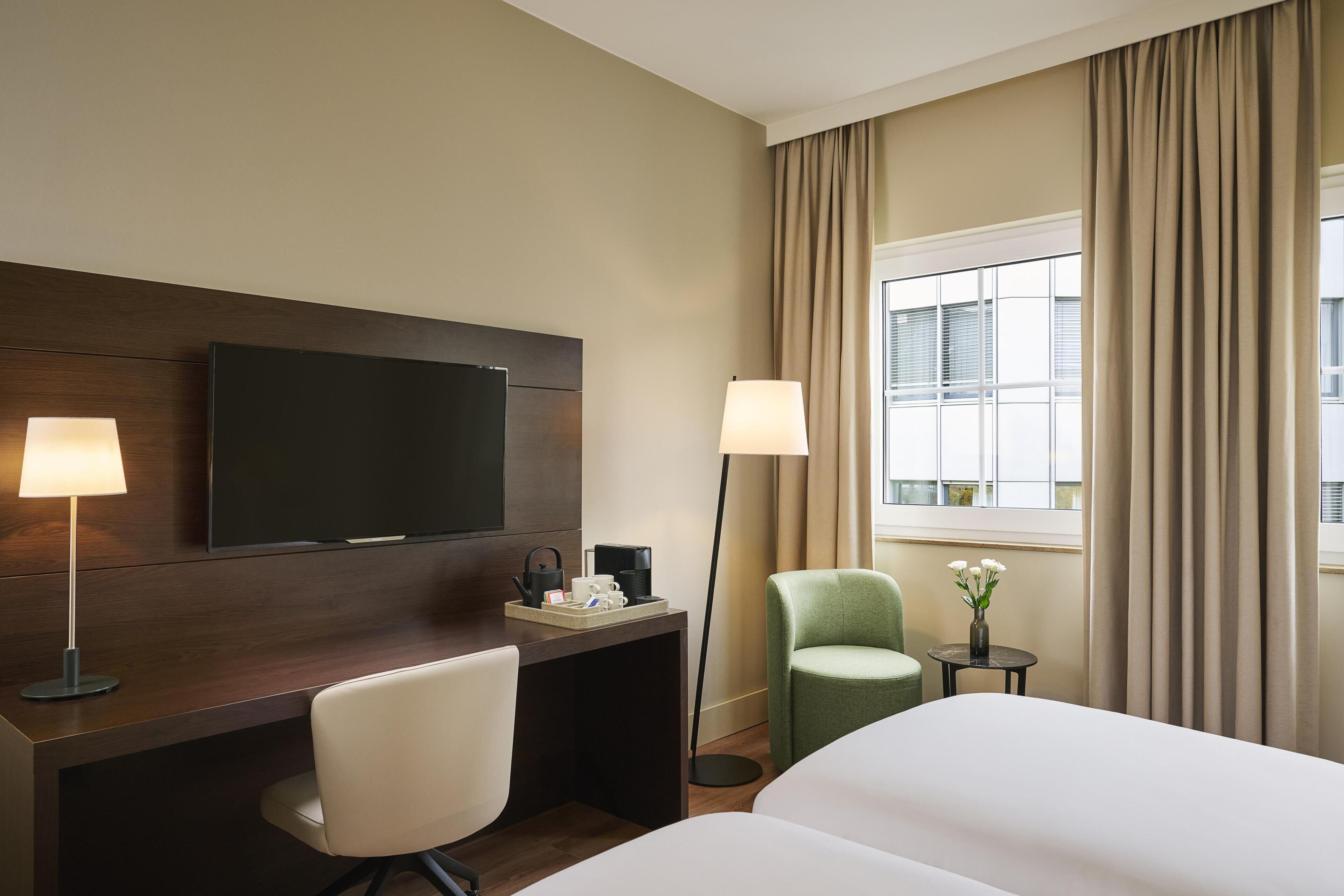 A hotel room with a bed, a desk with a chair, a television, a lamp, a small table with a vase of flowers, and a window with curtains.