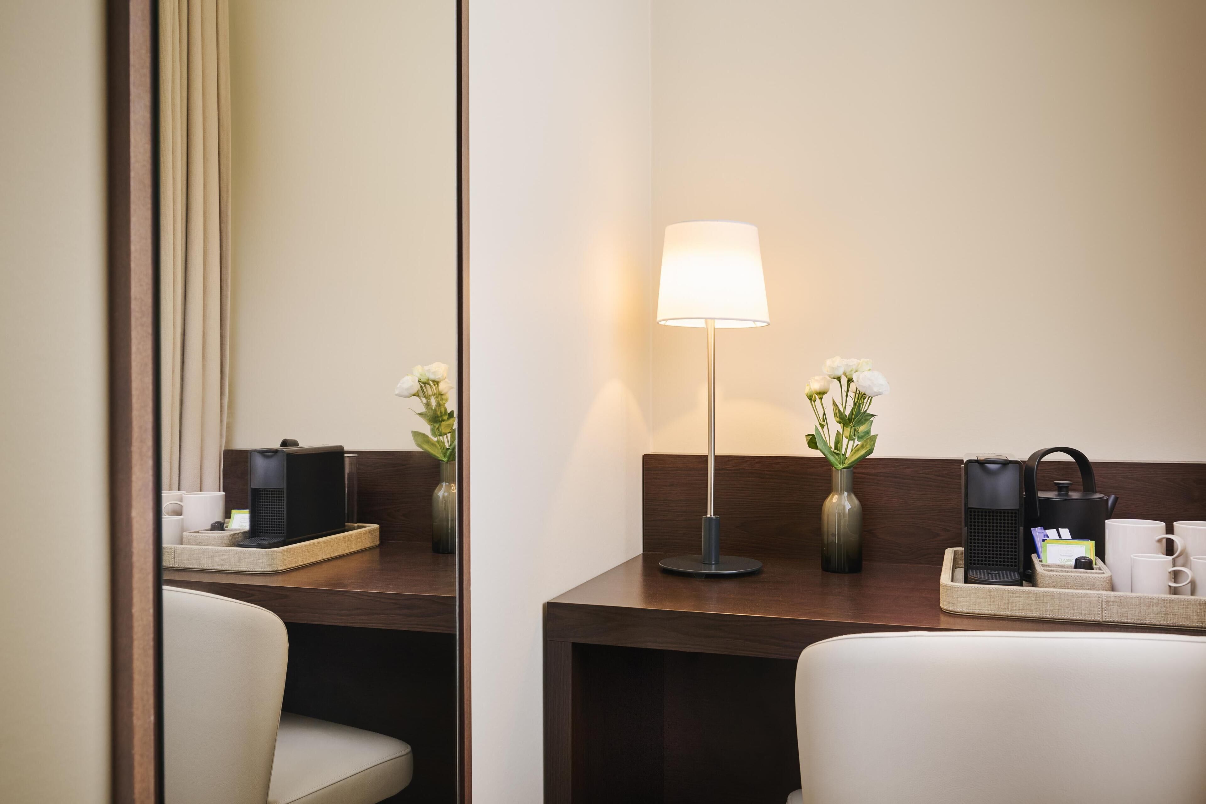 A hotel room with a wooden desk, a lamp, a vase with flowers, a tray with a kettle, and a coffee machine.