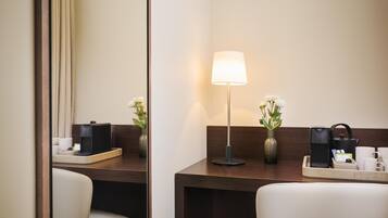 A hotel room with a wooden desk, a lamp, a vase with flowers, a tray with a kettle, and a coffee machine.